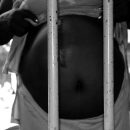 Foto em preto e branco de uma barriga de grávida encostada em uma grade de cela de cadeia