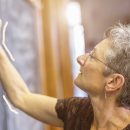 Imagem de uma professora atenta escrevendo na lousa. Ela tem cabelos grisalhos e usa óculos