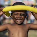 Imagem de um menino negro sorrindo com uma bexiga amarela em sua testa. Ele está com uma pintura preta no rosto