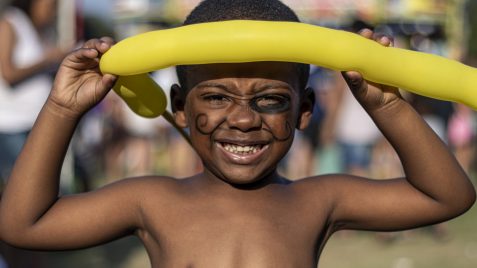 Imagem de um menino negro sorrindo com uma bexiga amarela em sua testa. Ele está com uma pintura preta no rosto