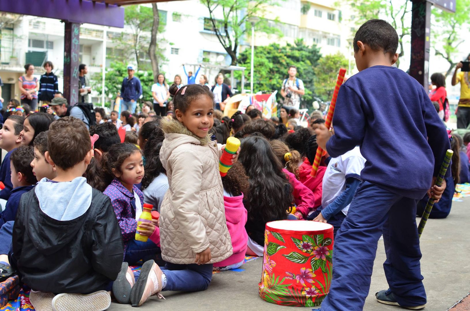 Foto de milhares de crianças sentadas no chão participando de ações coletivas. Uma menina está ajoelhada com um tambor colorido ao seu lado