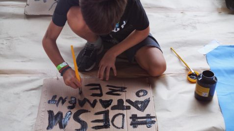Foto de um menino no chão fazendo um cartaz em um papelão com a palavra “desmatamento” em preto.