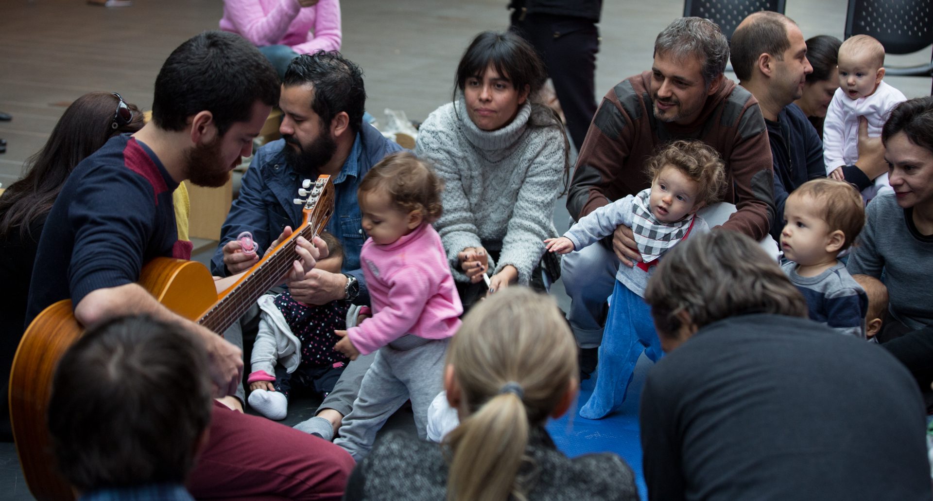 Do lado esquerdo da foto um homem de barba está tocando violão. Ele está sentado no chão assim como os bebês, mulheres e homens que estão diante dele. Alguns bebês estão de pé, sustentados pelos adultos e observam atentamente