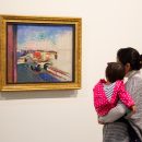 Foto de uma mulher em pé no museu segurando um bebê. Ambas estão olhando para um quadro cheio de texturas, formas e cores que está pendurado na parede.