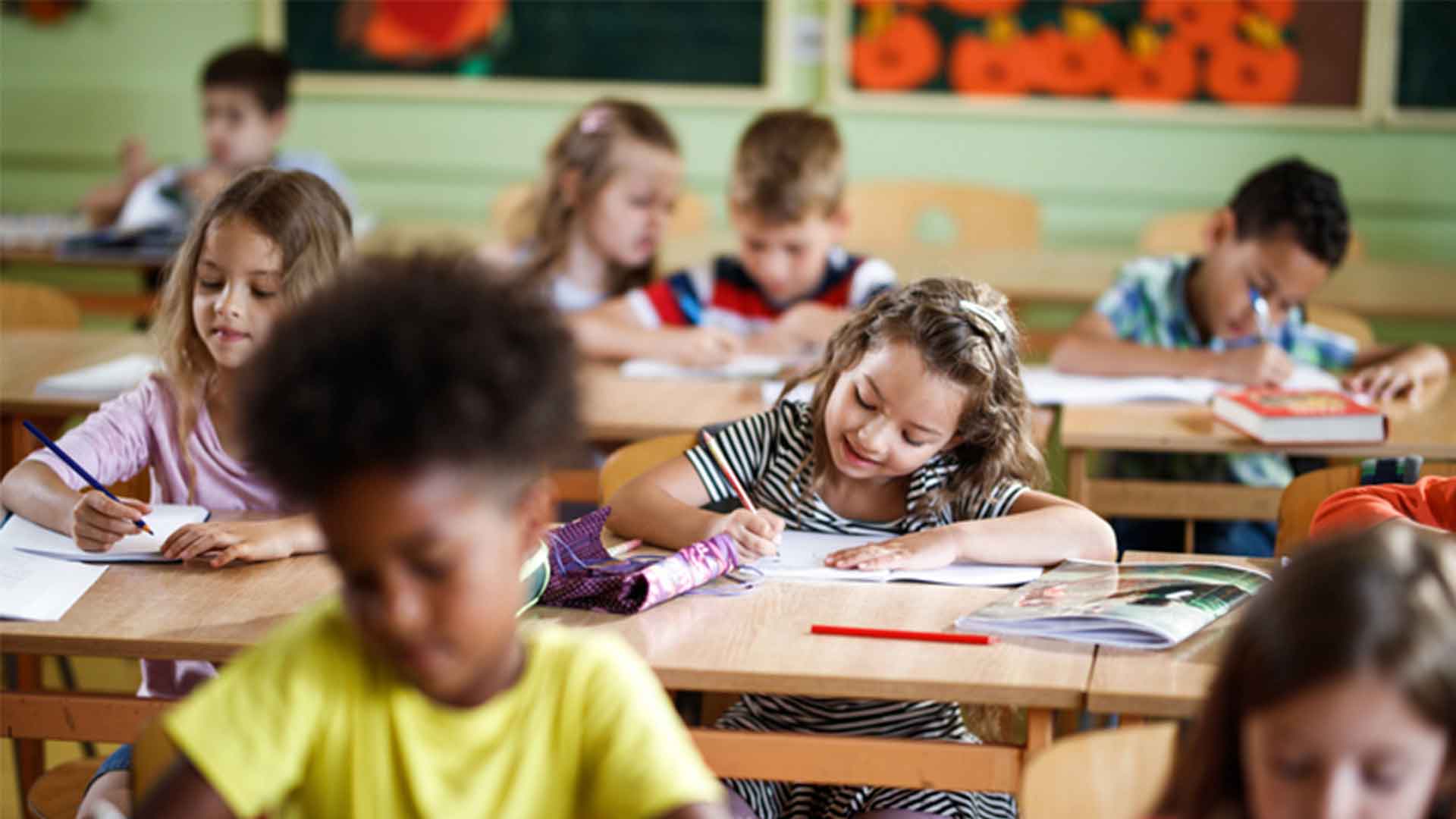 Imagem de uma menina sentada em sua carteira na escola sorrindo e desenhando. Em segundo plano aparece uma menina sentada a sua frente e atrás uma menina e um menino sentados na carteira da escola