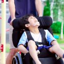 Imagem de uma criança com paralisia cerebral sentada em uma cadeira de rodas, e uma mulher levando a cadeira.