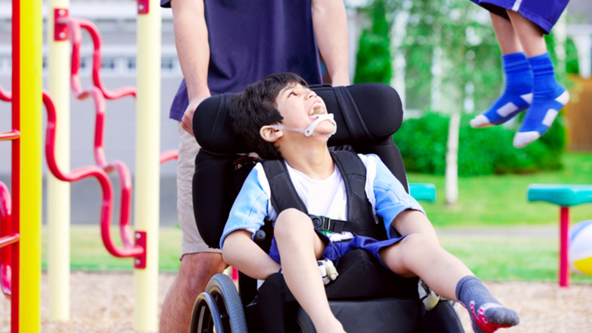 Imagem de uma criança com paralisia cerebral sentada em uma cadeira de rodas, e uma mulher levando a cadeira.