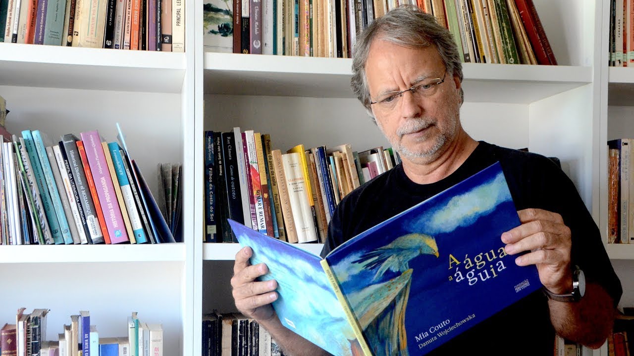 Imagem do escritor Mia Couto com seu livro “A água e a águia” em mãos. Ele está sentado lendo em frente a uma prateleira de livros.