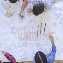 Imagem de uma visão aérea de um grupo multiétnico de crianças desenhando em um papel branco. As crianças estão sentadas em volta da mesa, criando um mural com canetas coloridas.