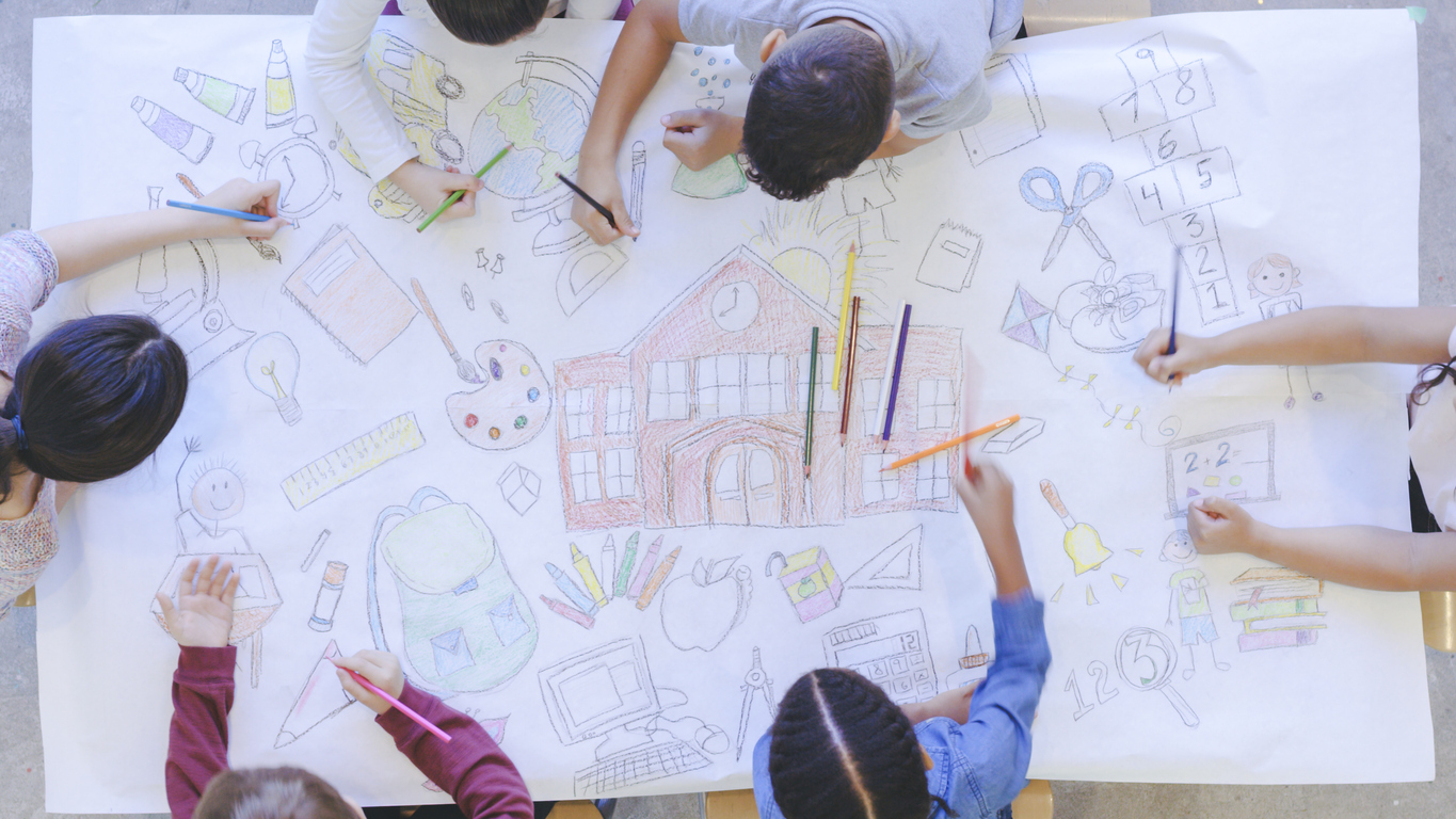 Imagem de uma visão aérea de um grupo multiétnico de crianças desenhando em um papel branco. As crianças estão sentadas em volta da mesa, criando um mural com canetas coloridas.