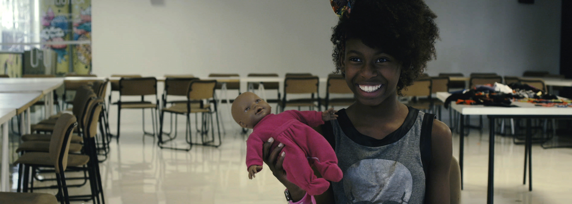 Capa do filme “Parece comigo” – Kelly Cristina Spinelli, foto de uma menina negra em sala de aula segurando uma boneca