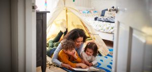 Imagem de um menino e uma menina com sua mãe deitados no chão do quarto. Eles estão lendo um livro embaixo de uma barraca armada no quarto infantil.