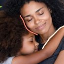 Foto de uma mãe e uma criança negras se abraçando com os olhos fechados. Artigo sobre a necessidade de cuidar de quem cuida.