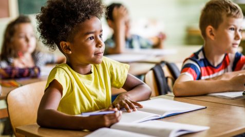 Imagem de uma menina negra sentada em uma mesa de sala de aula com livros escolares