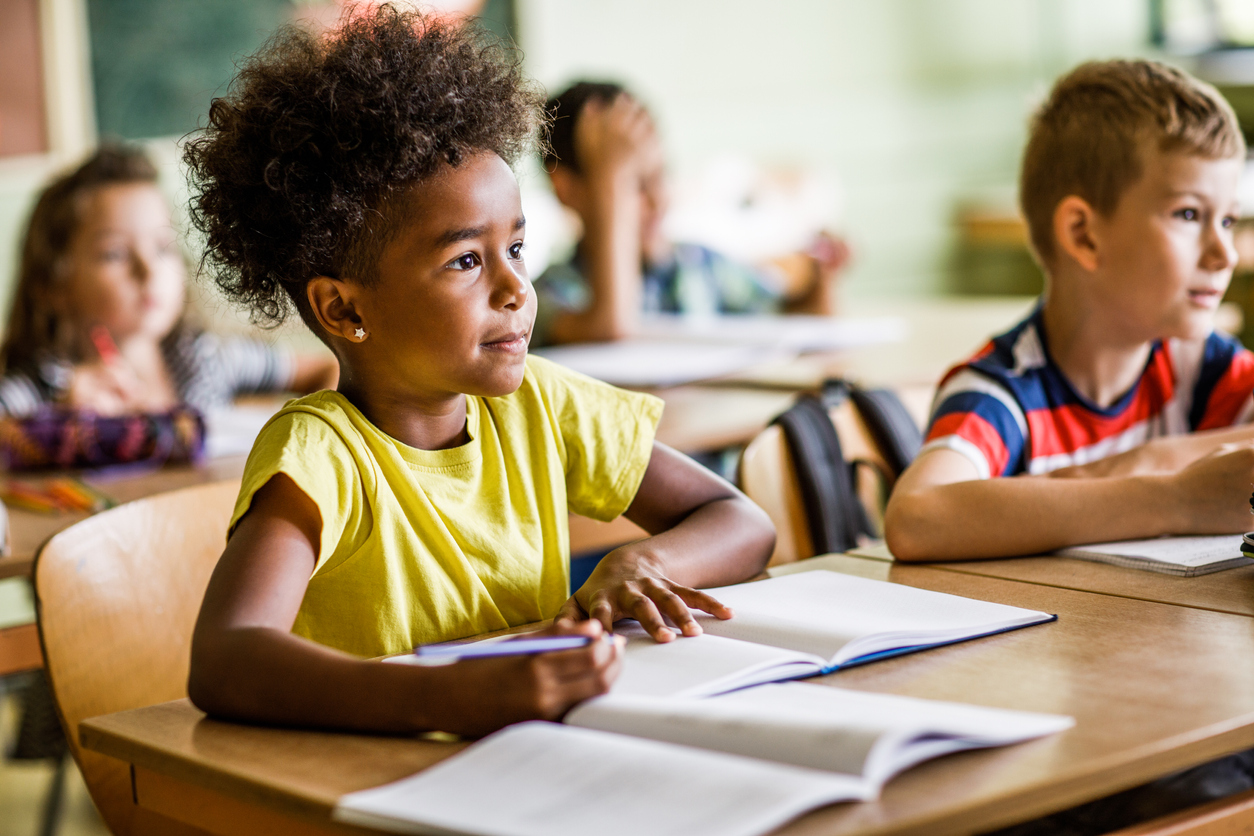 Imagem de uma menina negra sentada em uma mesa de sala de aula com livros escolares