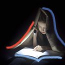 Imagem em preto e branco de uma menina lendo um livro com uma coberta na cabeça, e uma lanterna em suas mãos iluminando o livro