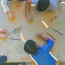 Imagem de quatro crianças rabiscando um papel pardo, e desenhando o planeta terra, um satélite, um carro movido a energia elétrica, projeções positivas de futuro.