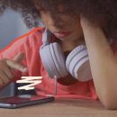 Imagem de uma menina negra, aparentando 12 anos, com cabelos encaracolados e vestindo uma camiseta da cor rosa, está sentada à mesa e mexendo no celular.Tem um fone branco em volta do pescoço.
