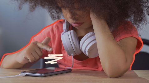 Imagem de uma menina negra, aparentando 12 anos, com cabelos encaracolados e vestindo uma camiseta da cor rosa, está sentada à mesa e mexendo no celular.Tem um fone branco em volta do pescoço.