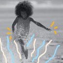 Foto de um menino com cabelo black power. Ele corre feliz pela água que bate na areia da praia