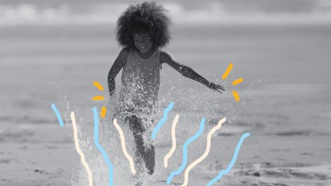 Foto de um menino com cabelo black power. Ele corre feliz pela água que bate na areia da praia