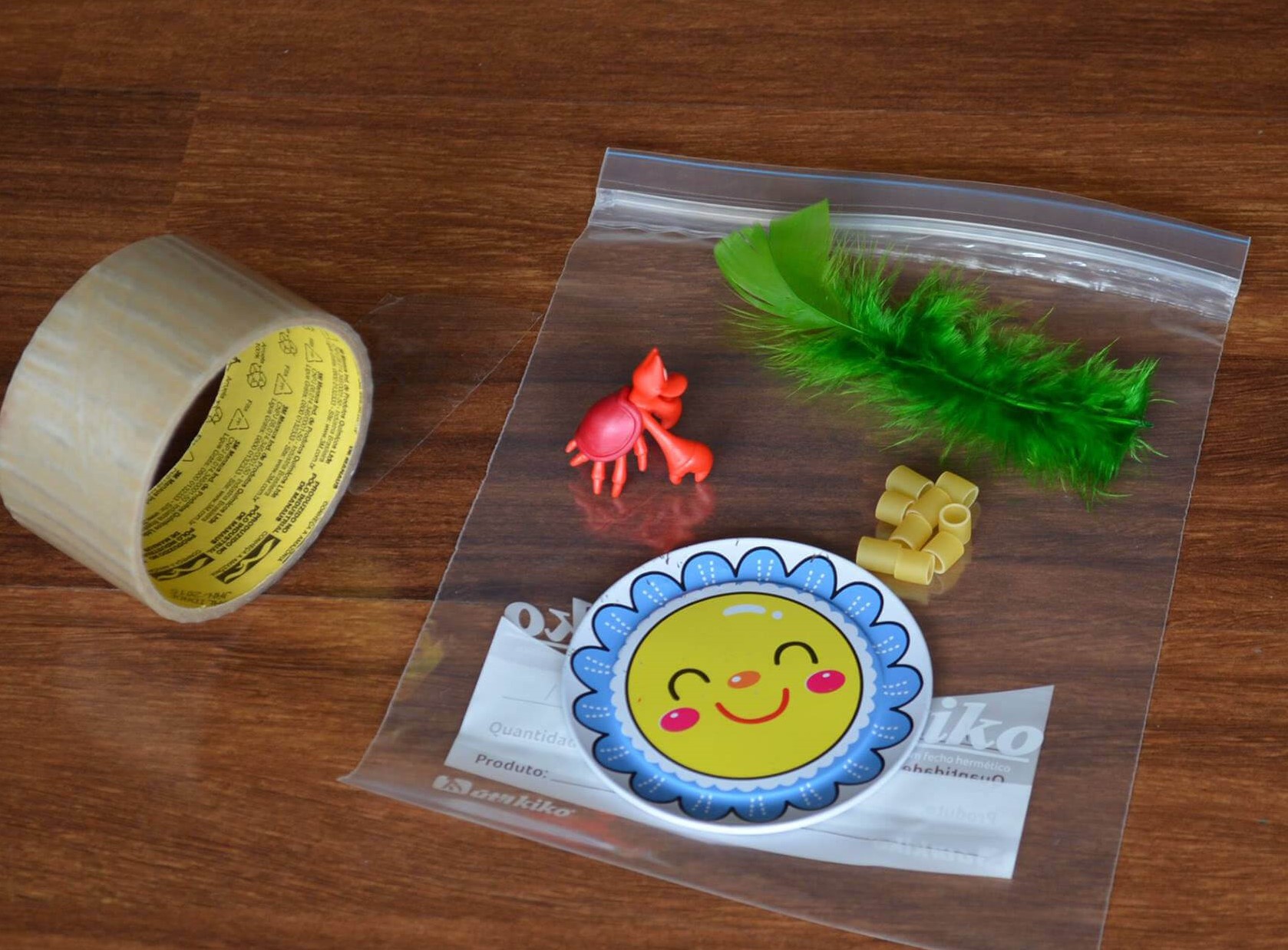 Foto de um saco plástico, com uma pena, pequenas peças quadras e um caranguejo de plástico. Ao lado, uma fita adesiva