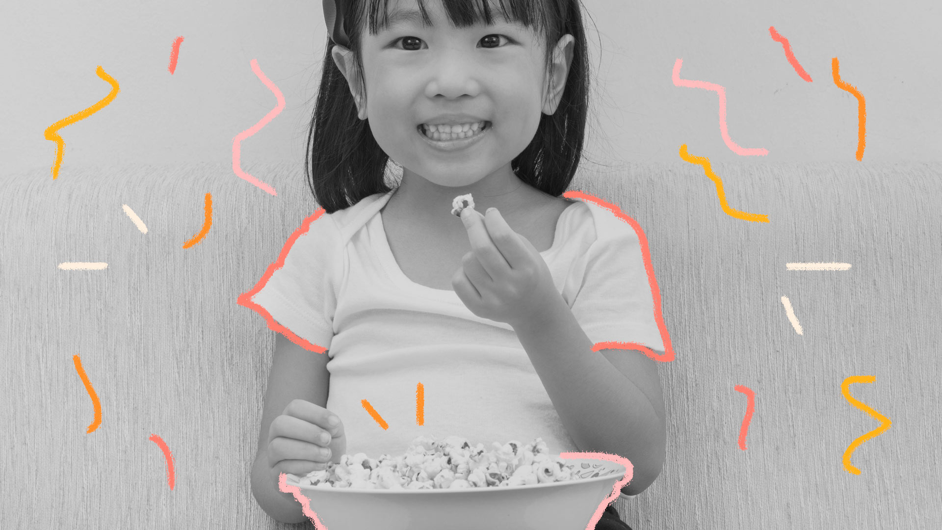 Menina asiática segura uma bacia de pipoca no colo, como se estivesse assistindo a um filme infantil, e sorri para a câmera