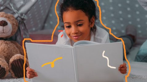 Imagem de uma menina negra aparentando ter 6 anos de idade sorri enquanto lê um livro infantil