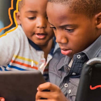 Imagem de dois irmãos negros aparecem na foto olhando para o mesmo tablet. O menino mais velho é cadeirante.
