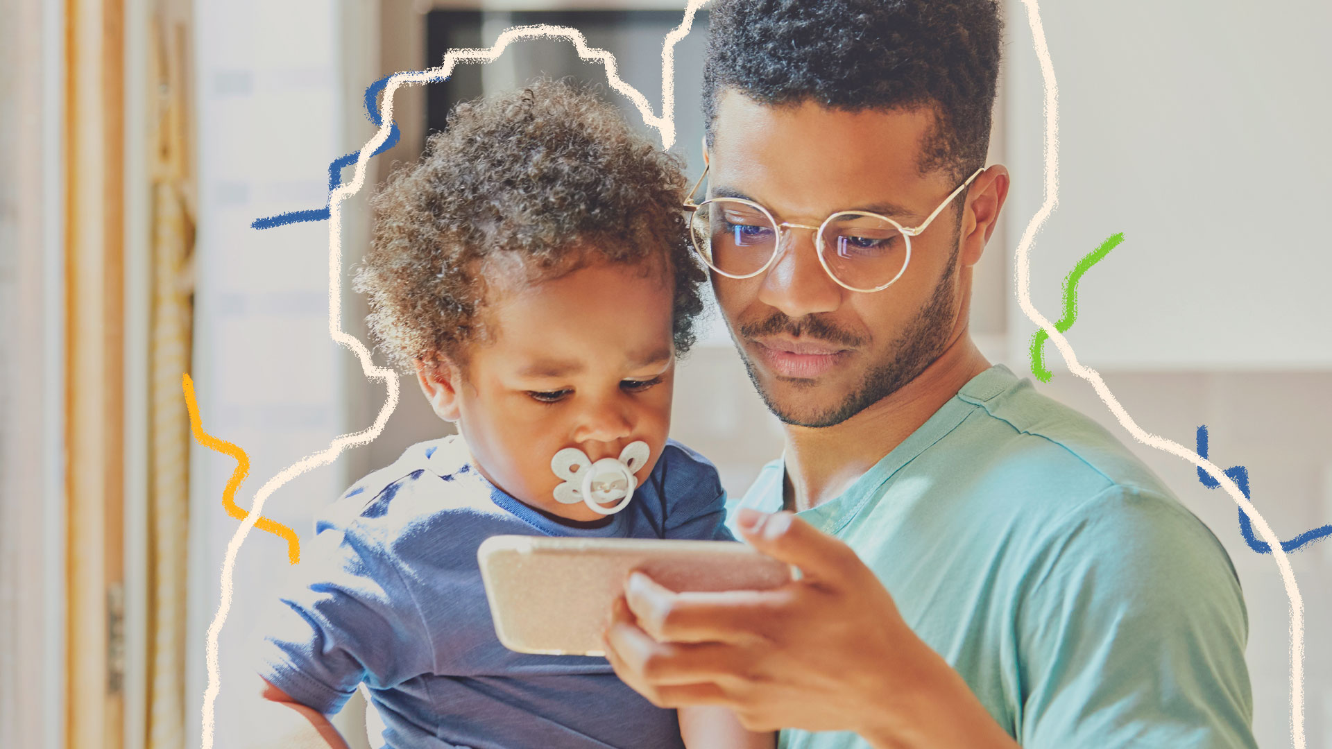 Criança e internet: pai segura bebê no colo e ambos olham para a tela do celular. O menino está com chupeta na boca e o adulto usa óculos. Os dois são negros.