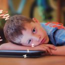 Imagem de um menino de cabelo castanho aparece debruçado sobre um tablet e aparenta estar um pouco cansado. Texto sobre a influência das telas no desenvolvimento da criança