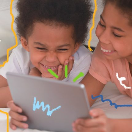 Imagem de duas crianças negras estão vendo algum conteúdo em um tablet e sorrindo.
