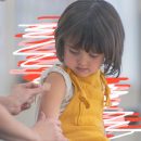 Vacinação em crianças: uma menininha morena de cabelos curtos aparece na foto com um macacão amarelo e a blusinha branca com a manga arregaçada. Isso porque ela acaba de receber uma injeção. A profissional de saúde, de quem aparece apenas as mãos, está lhe colocando um curativo.