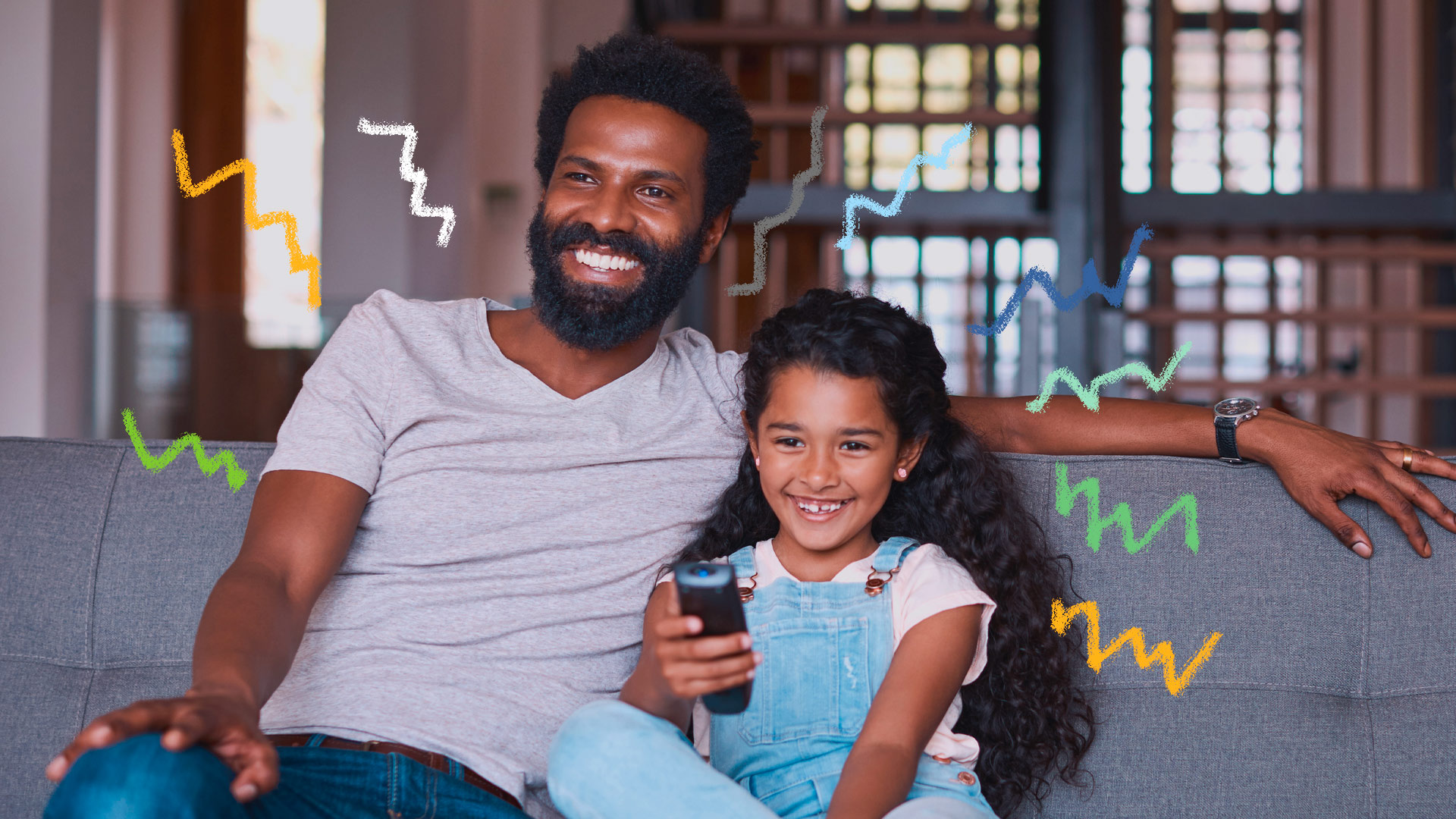 Foto de pai e filha, ambos negros, estão sentados em um sofá cinza. Ela segura um controle remoto e aponta para a televisão. No fundo, uma janela. Os dois vestem blusa branca e peças em jeans e sorriem. Lista com representações de pais em animações infantis