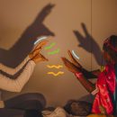 Imagem de mãe e filha, ambas negras, estão brincando de sombras projetadas na parede de um quarto e rindo