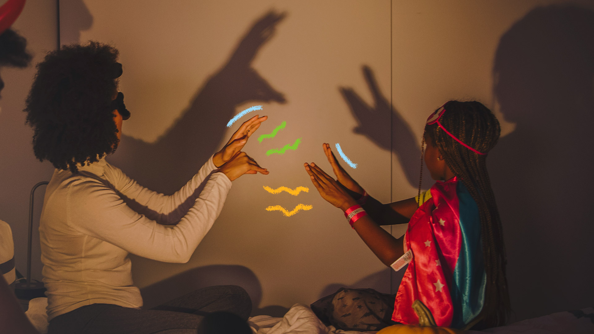 Imagem de mãe e filha, ambas negras, estão brincando de sombras projetadas na parede de um quarto e rindo