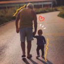 Foto de um avô dando as mãos ao neto bebê acompanhando-o nos primeiros passos e ambos caminham lado a lado, de costas, seguindo por uma estrada