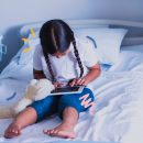 Imagem de uma menina com duas tranças, acompanhada de um ursinho de pelúcia. Ela está sentada em uma cama de hospital e apoia no colo um tablet onde deve acessar histórias infantis.