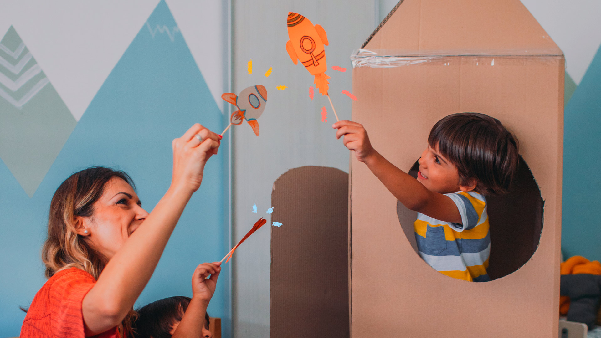 Imagem de mãe e filho brincando com elementos simples de papel e papelão, fazendo pequenos e grandes foguetes.
