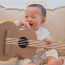 Imagem de um bebê sentado no sofá sorrindo, com um violão feito de caixa de papel