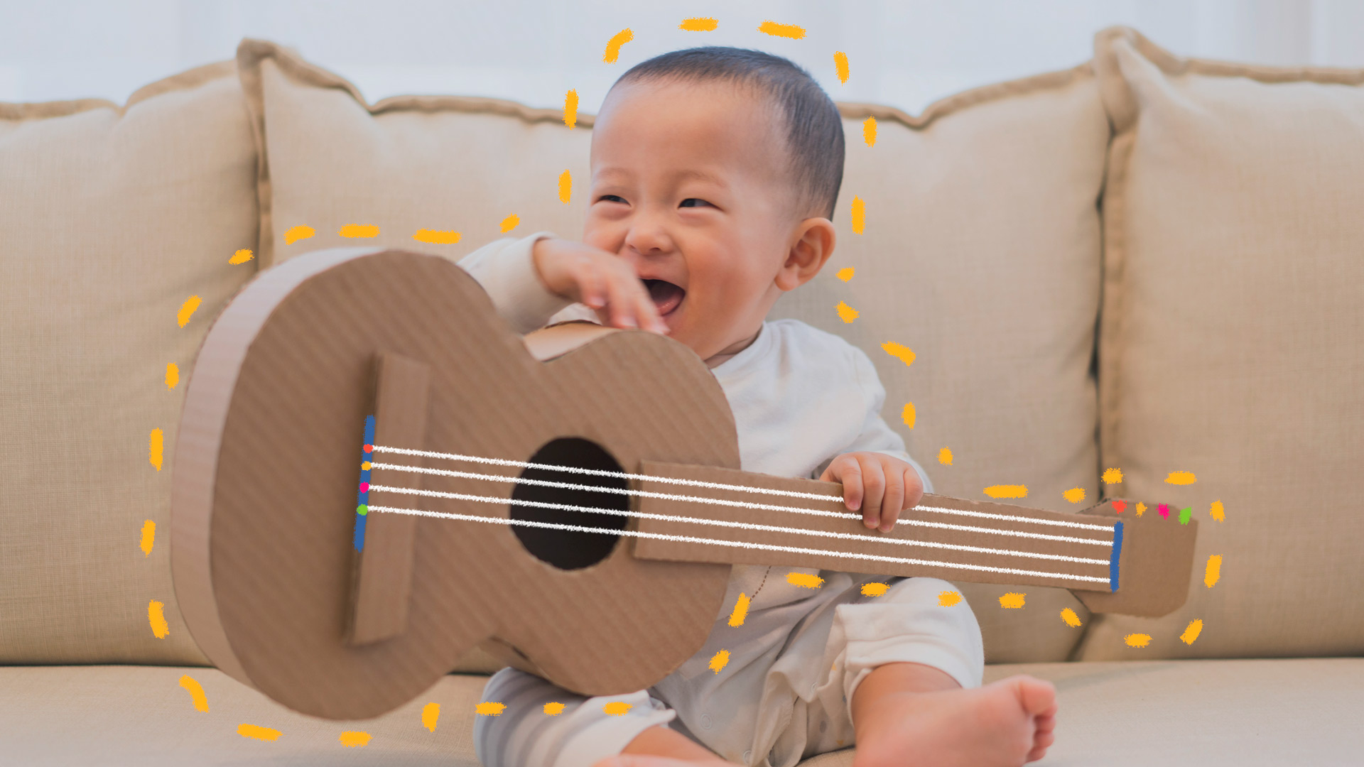 Imagem de um bebê sentado no sofá sorrindo, com um violão feito de caixa de papel