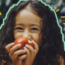 Imagem de uma menina segurando uma maçã mordida em frente ao rosto e sorri. A imagem ilustra uma entrevista com Gabriela Kapim