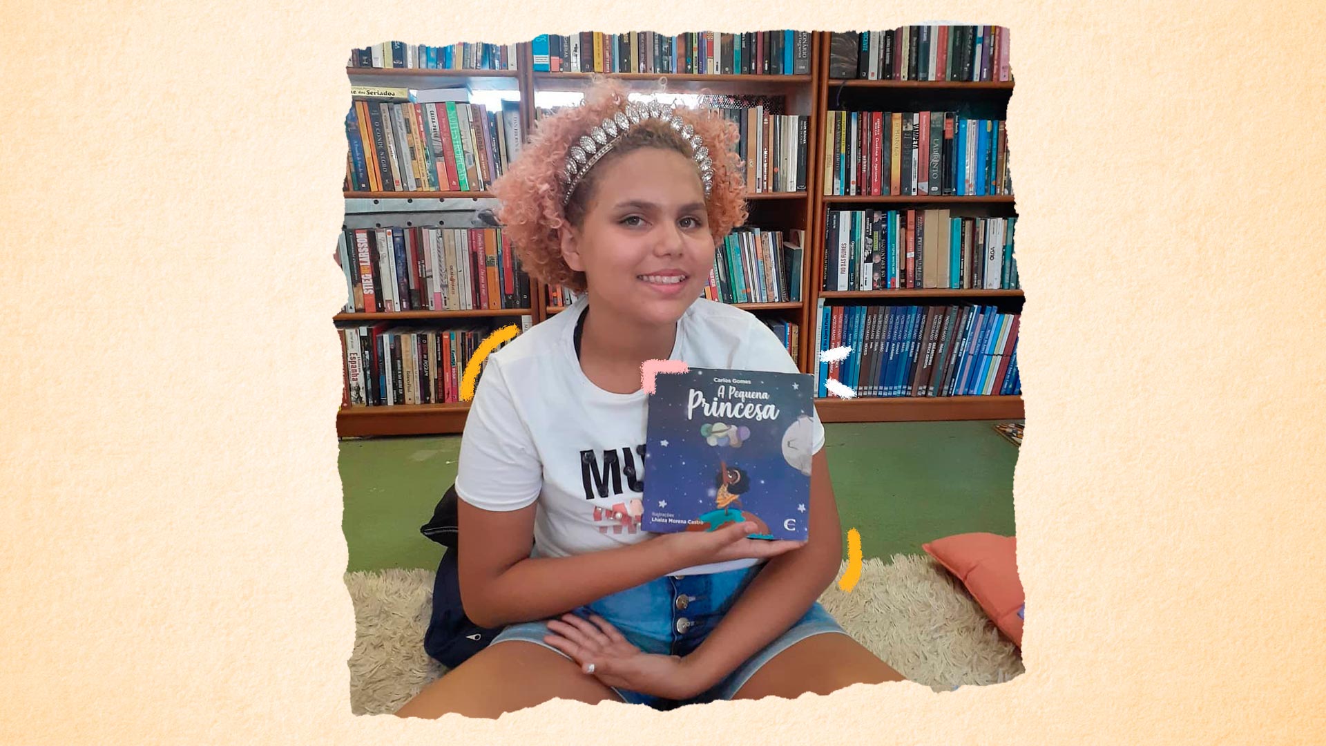 Imagem de Lua Oliveira sentada no chão da biblioteca segurando o livro "A pequena princesa", dedicado a ela.