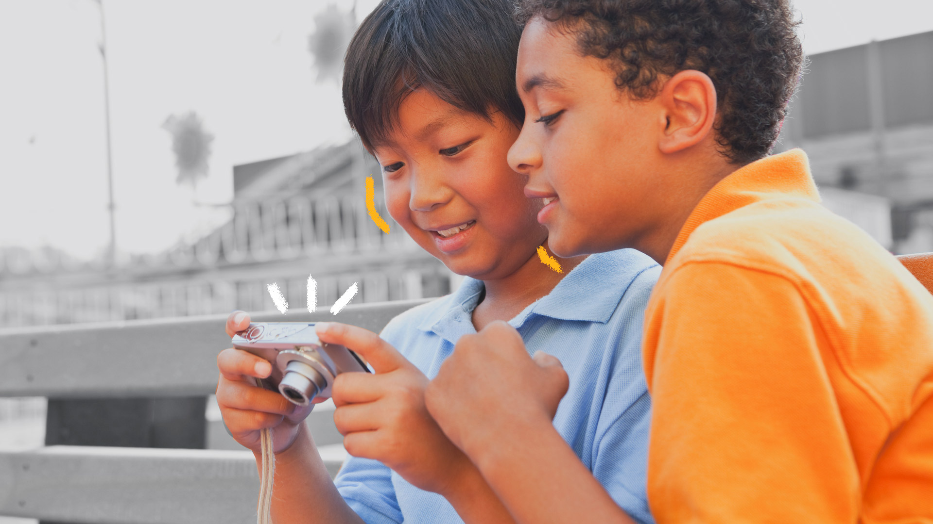 Imagem de dois meninos, um negro de camiseta laranja e um asiático de camiseta azul, olham através de uma câmera digital