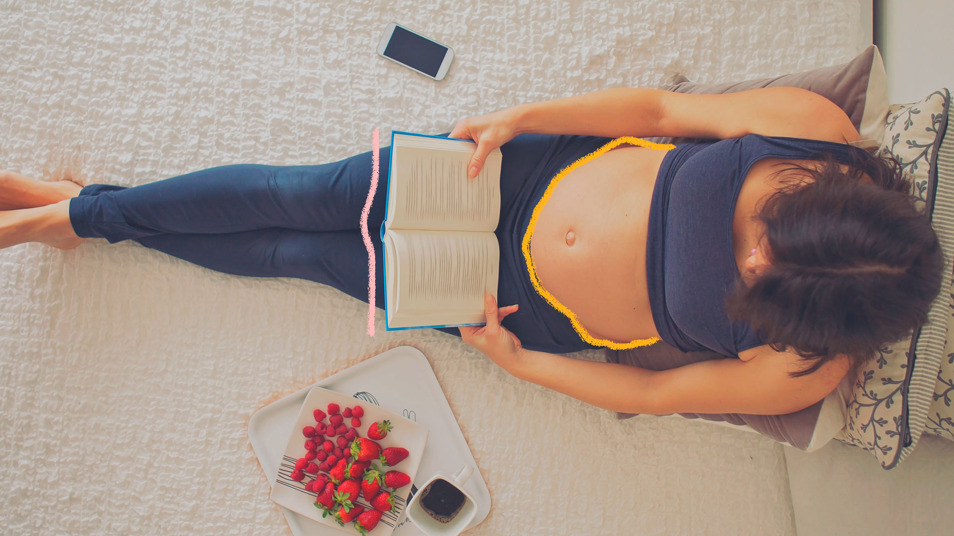 Imagem de uma mulher grávida lendo um livro sentada no tapete. Ao seu lado, um celular, frutas e café.