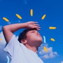 Imagem de um menino negro vestindo uma camiseta branca. Ele está com a mão direita cobrindo acima da cabeça, fazendo o gesto de olhar ao alto e horizonte. No fundo, um céu azul.