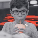 Imagem em preto e branco de um menino, de óculos, tomando uma bebida em um copo plástico com canudinho.