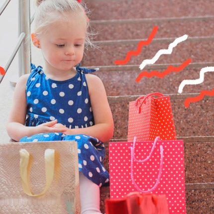 Imagem de uma menina com vestido azul de bolinhas brancas sentada na escada olha para sacolas de compras à sua volta