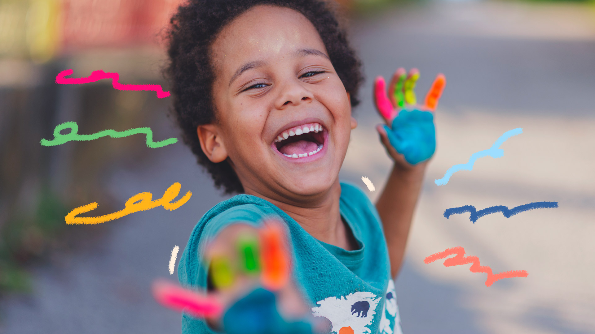 Imagem de um menino sorrindo e mostrando as duas mãos coloridas com tinta cada dedo com uma cor