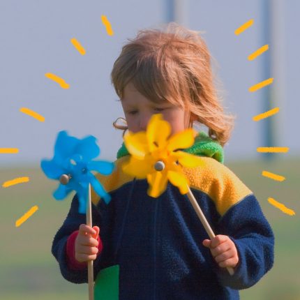 Imagem de um bebê segurando dois cataventos, um azul e outro amarelo, em um campo aberto.
